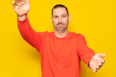 You did it. Congratulations. Happy cheerful positive hispanic man with beard standing over yellow colored background looking at camera, smiling and opening arms for warm hug.の写真素材