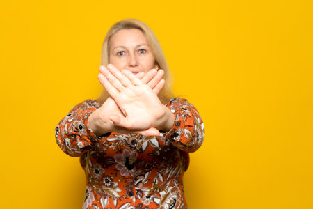 Pretty blonde Caucasian woman in her 40s wearing a patterned sweater, making the prohibition gesture with crossed palms in front of the camera, isolated over yellow studio background.の写真素材