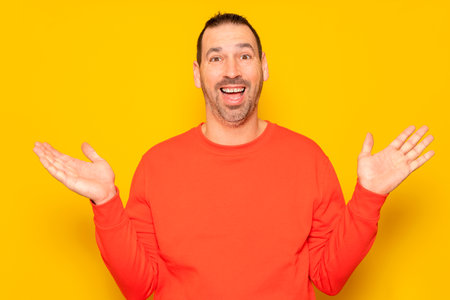 Funny smiling man in red sweater showing palms up, unexpected surprise gesture isolated over yellow background. Attractive happy man listening to good news, catching somethingの写真素材
