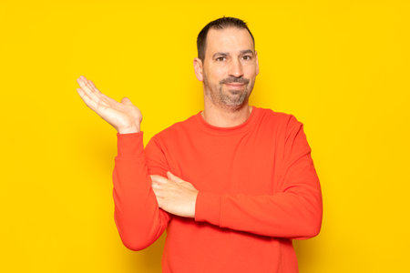 Hispanic man with a beard and red sweater in a friendly attitude offering something on the palm of his hand, isolated on a yellow backgroundの写真素材
