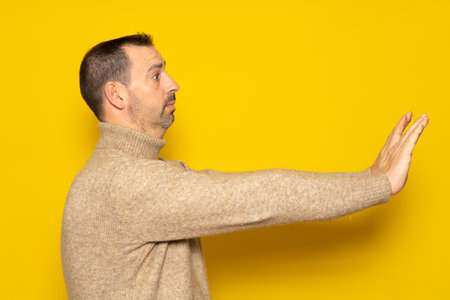 Side view of a scared man standing with a scared or worried face, looking at the camera and blocking with his hands. Indoor studio shot isolated on yellow backgroundの写真素材