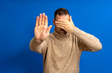 Man in his 40s with a beard wearing a turtleneck covering his eyes with his hands and making a stop gesture with an expression of sadness and fear. Embarrassed and negative concept.の写真素材