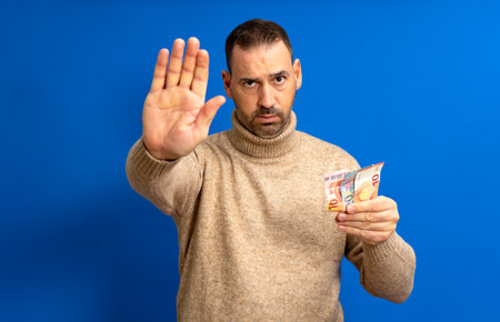 Bearded hispanic man in his 40s wearing a beige turtleneck holding several euro bills and making the stop sign isolated over blue backgroundの写真素材