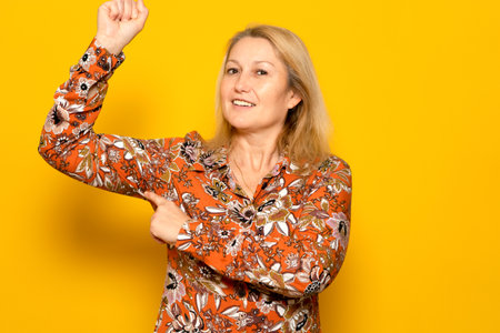Strong independent caucasian woman with blond hair showing her arm muscle and point it with finger, proud of her strength and leadership skills, emancipation. Indoor isolated on yellow backgroundの写真素材