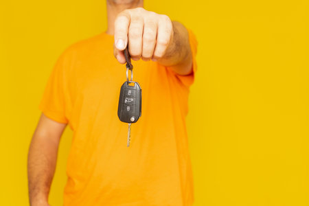 Unidentified man stretching out his arm and offering a car key, isolated on yellow studio backgroundの写真素材