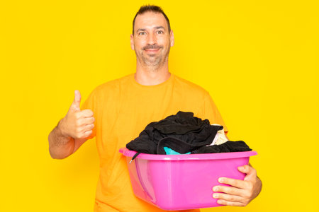 Bearded hispanic man doing housework holding plastic basket with clothes raising thumb up, excellent symbol. Isolated on yellow background.の写真素材