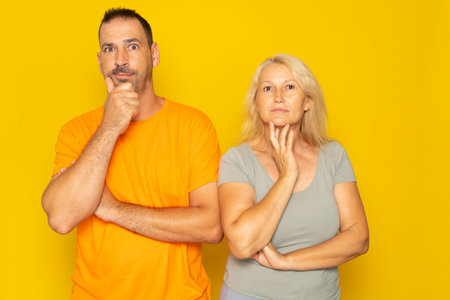 Caucasian real couple in thoughtful pose with hand on chin making a difficult decision, isolated on yellow studio background.の写真素材