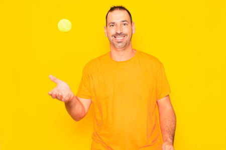 Hispanic man with beard in his 40s throwing a tennis ball in the air isolated on yellow studio background.の写真素材