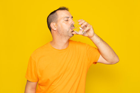 Hispanic man with beard in his 40s drinking fresh water in a glass glass, isolated on yellow background.の写真素材