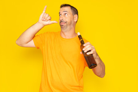 Hispanic man with beard in his 40s making a go drunk gesture with a bottle of wine in his hand, isolated on yellow studio backgroundの写真素材