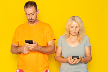 Middle aged couple looking at their cell phone ignoring each other isolated on yellow studio background. Concept of social distancing due to technologyの写真素材