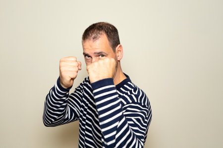 Bearded Hispanic man in his 40s wearing a striped sweater with his fists in defense position prepared to fight, isolated on beige isolated backgroundの写真素材