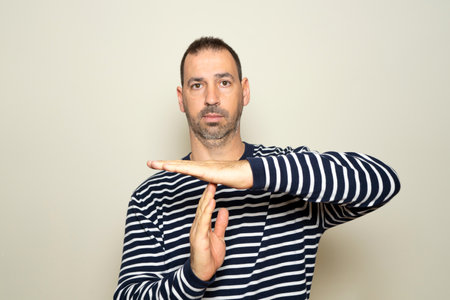 Bearded Hispanic man in his 40s wearing a striped sweater calling for a time out with his hands isolated on a beige background, he needs a break from the fast pace of his lifeの写真素材