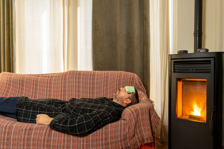 Sick Hispanic man lying on the couch with a robe and a cloth on his forehead in the heat of a pellet stoveの写真素材