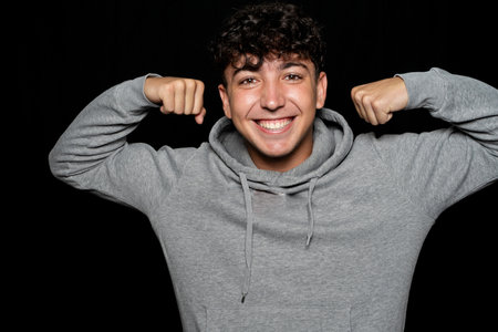 Young man with curly hair about 25 years old wearing sports sweatshirt making muscle gesture over dark backgroundの写真素材