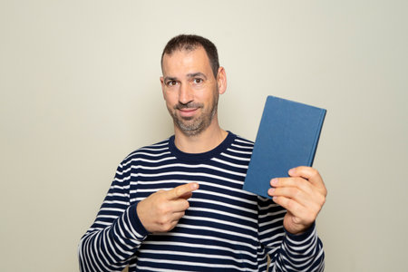 Bearded Hispanic man in his 40s pointing at a paper book on a beige background, making a recommendation for traditional reading over digital readingの写真素材