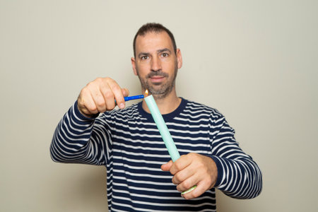 Bearded hispanic man in his 40s dressed in a striped sweater lighting an elongated wax candle with a lighter, isolated over beige background. Selective focus on the candleの写真素材