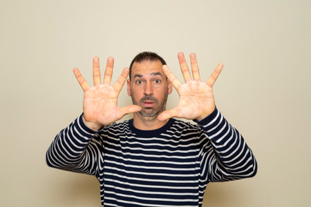 Bearded Hispanic man in his forties wearing a striped sweater feels dumbfounded and scared, fearing something scary, with open hands in front. Isolated over beige studio background.の写真素材