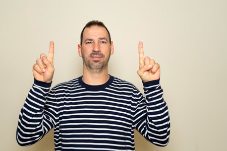 Bearded hispanic man around 40s wearing striped sweater smiling with hands pointing to empty space above, isolated over beige background.の写真素材
