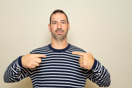 Bearded Hispanic man in his 40s wearing a striped sweater pointing at himself proud of the job he just did.の写真素材