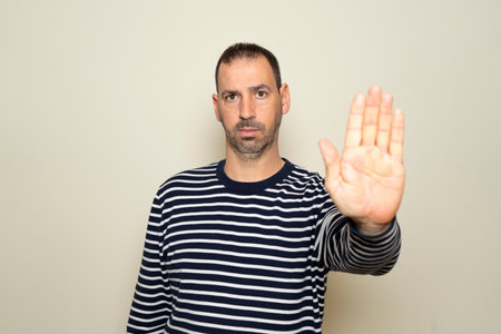 Portrait of a bearded Hispanic man in his 40s wearing a striped sweater and making a stop gesture with his palm. Isolated on beige backgroundの写真素材