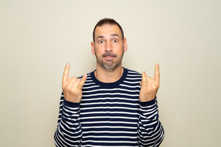 Portrait of bearded man showing rock and roll gesture, heavy metal sign, exclaiming with joy. Indoor studio photo isolated on beige background.の写真素材