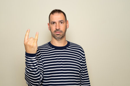 Portrait of bearded man showing rock and roll gesture, heavy metal sign, exclaiming with joy. Indoor studio photo isolated on beige background.の写真素材