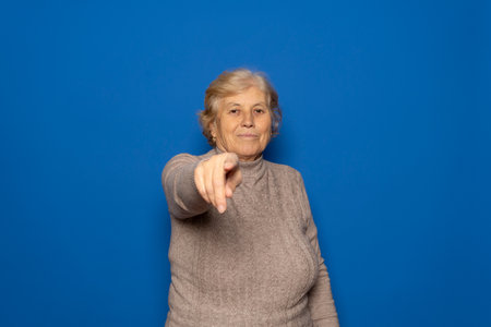 Caucasian woman in her 70s wearing a dark beige sweater pointing at the camera while smiling kindly, isolated over blue studio background.の写真素材