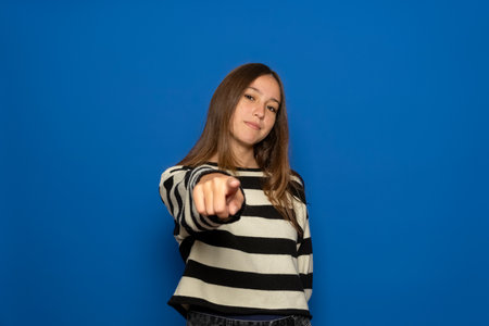 Hispanic teenage girl about 15 years old wearing a striped sweater pointing at the camera with a satisfied, confident and friendly smile. Isolated over blue studio backgroundの写真素材