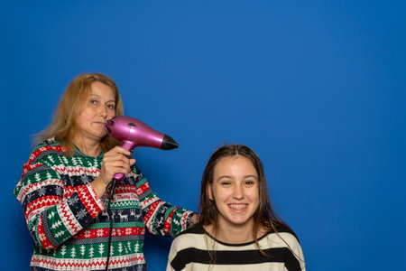 Caucasian woman in her 40s blow-drying her teenage daughters hair, isolated on blue background.の写真素材