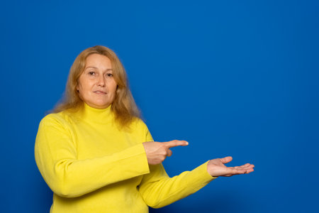Caucasian woman in her 40s wearing a yellow sweater pointing with her finger and selecting a product isolated on a blue background.の写真素材