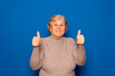 Caucasian mature woman about 70 years old wearing dark beige sweater with thumbs up in sign of...