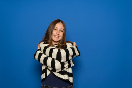 Girl about 15 years old with braces wearing striped sweater smiling and hugging herself isolated over vibrant blue color background.の写真素材