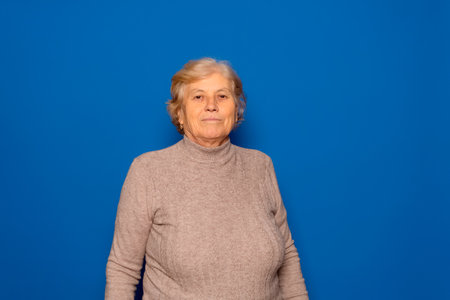 Horizontal portrait of a Caucasian woman in her 70s posing proudly and satisfied with the stage of her life she is living, isolated on a blue studio backgroundの写真素材