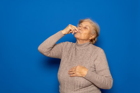 Caucasian woman in her 70s wearing a dark beige sweater licking her fingers after tasting a delicacy. Isolated on blue background.の写真素材