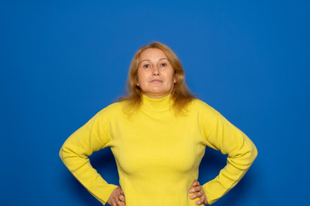 Caucasian woman in her 40s wearing yellow sweater posing happy with arms on hips and looking at camera over blue background with copy space. Confident woman is happy and feeling relaxedの写真素材