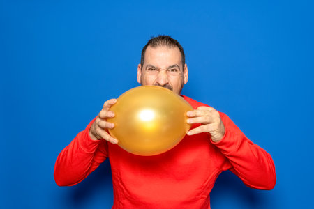 Bearded man in his 40s wearing a red sweater holding an orange balloon annoyed and frustrated with the celebrations, isolated on blue studio backgroundの写真素材