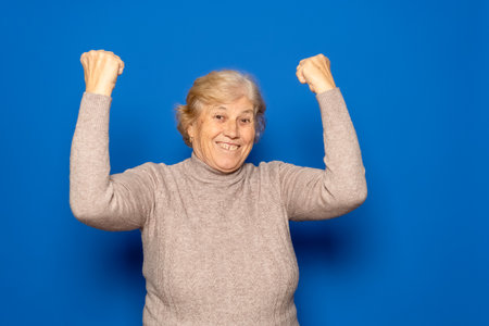 Caucasian woman in her 70s wearing a dark beige sweater celebrating happily with her fists raised, isolated on blue studio background.の写真素材