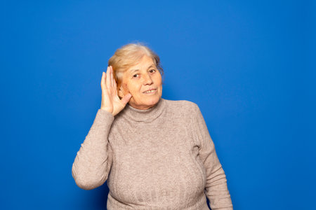 Caucasian woman in her 70s wearing a dark beige sweater putting her hand to her ear because she cant hear the most interesting gossip, isolated on blue backgroundの写真素材
