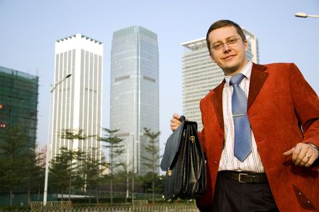 Businessman in modern city, wearing red suit and blue tie, holding briefcase in his hand. Modern skyscrapers as background.の写真素材