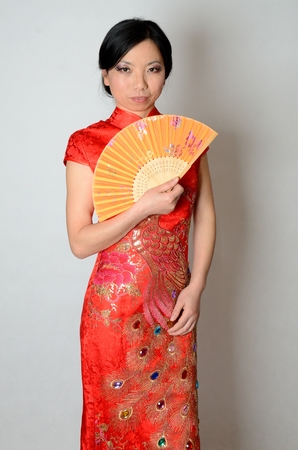 Elegant Chinese lady wearing traditional red dress. Pretty Asian female model holding fan in her hand. の写真素材