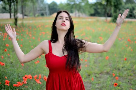 Female model with red dress. Girl meditating in open field, meadow with poppies in background. Model with closed eyes and open arms.の写真素材