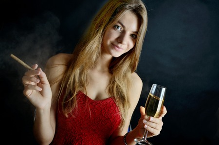 Young female sits with cigarillo and glas of white wine in her hand. Woman in red evening dress.の写真素材