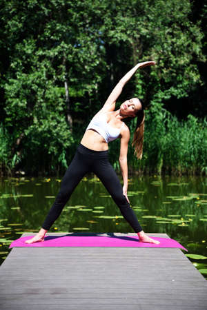 Woman in her 30's practicing yoga and meditation. Fit woman on wooden platform at the pond, surrounded by trees and water.の写真素材