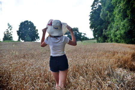 Outdoor photos of young woman in her 30's. Summer season in Poland. Woman with straw hat stands in wheat's field.の写真素材