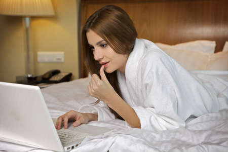 Beautyful brunette woman looking at a laptop lying on bed at homeの写真素材