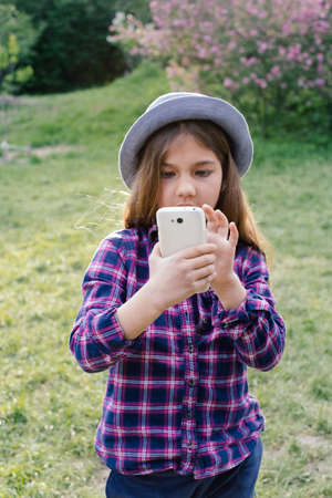 Adorable pre-teen tweenie brunette kid girl with her smartphone in the spring park. Dressed in shirt and blue hatの写真素材