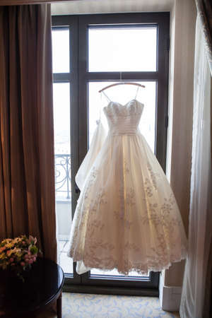 Bright beige wedding dress hanging on a hanger near a windowの写真素材
