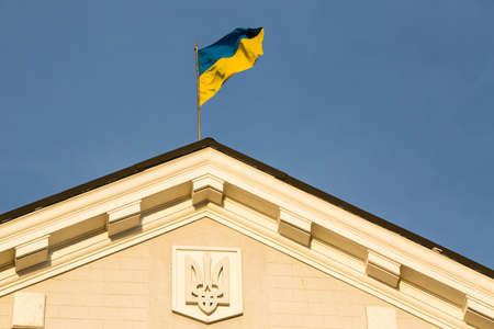 Ukrainian flag on top of government building with Ukrainian coat of arms trident at blue sky...