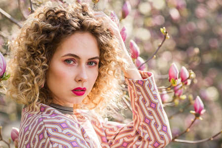 Closeup portrait of blondy beautiful girl with curly long hair. Woman walks in the garden of blooming pink magnoliaの写真素材